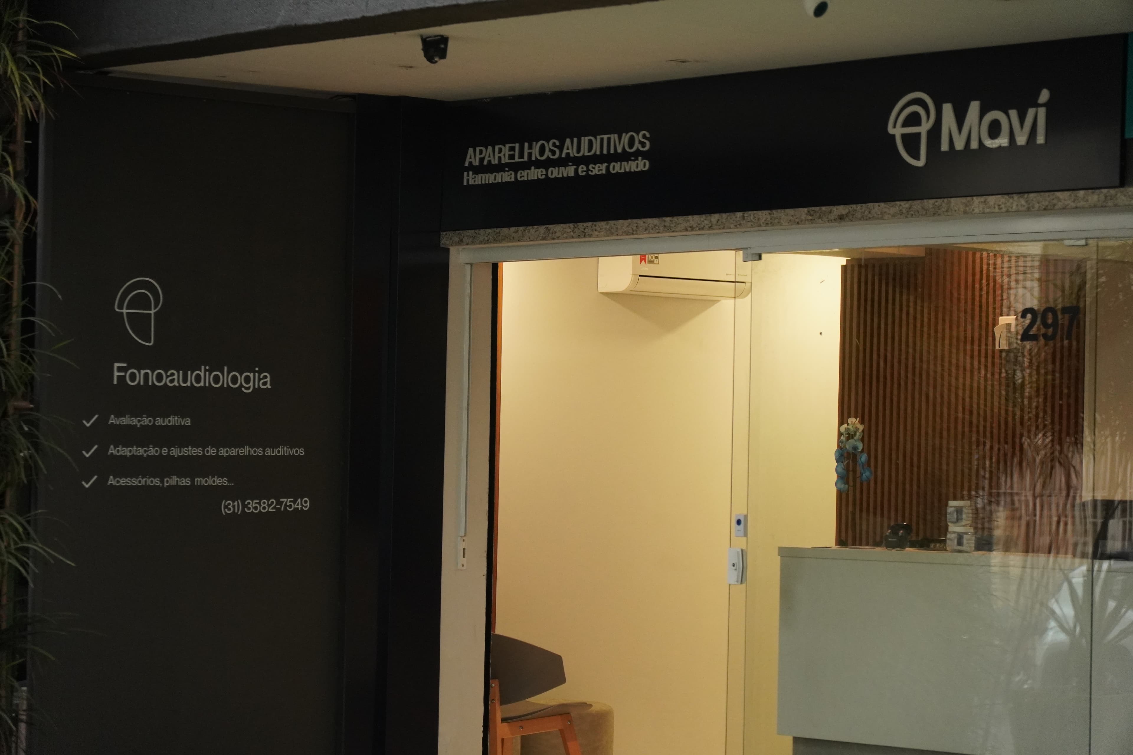 Fachada da Maví, centro auditivo em Belo Horizonte, com a placa "Aparelhos Auditivos: Harmonia entre ouvir e ser ouvido" e a entrada iluminada da loja